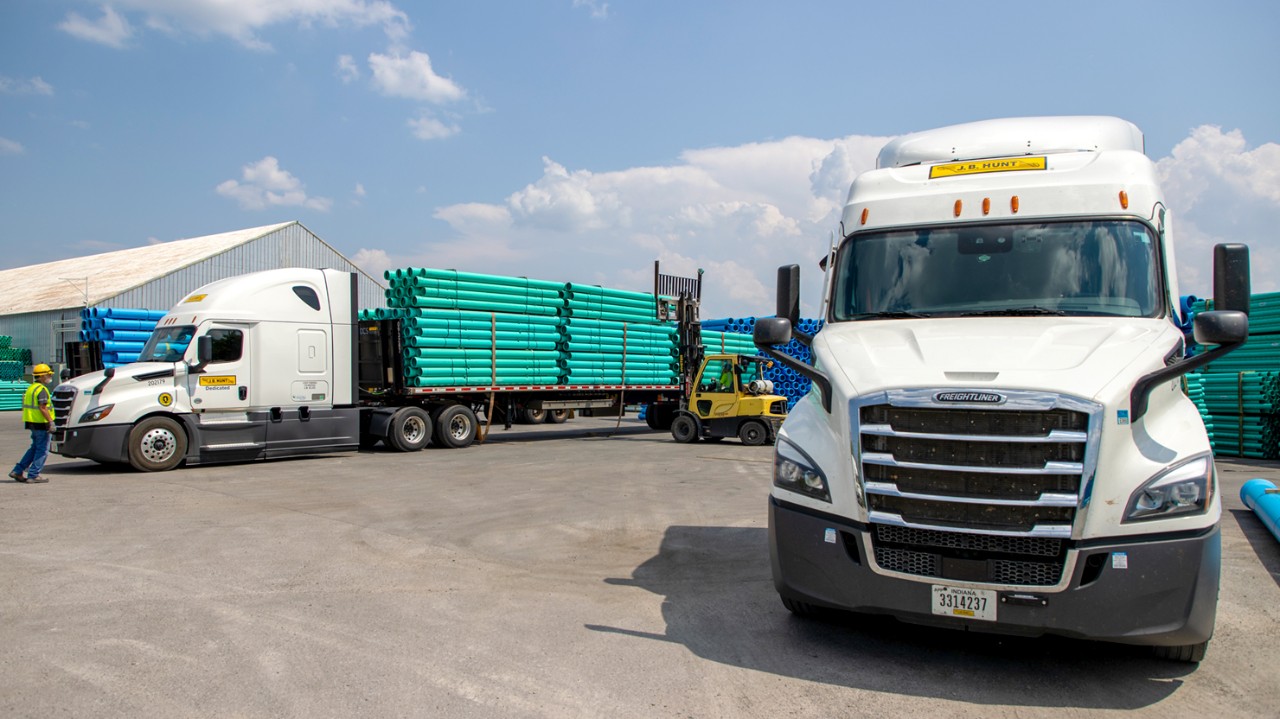 Two J.B. Hunt Freightliner semi-trucks parked in an industrial yard, each carrying flatbed trailers loaded with neatly stacked green pipes. A yellow forklift is in the process of loading or unloading pipes between the trucks, while a worker in a yellow safety vest and hard hat walks nearby. A large warehouse is visible in the background under a partly cloudy sky.