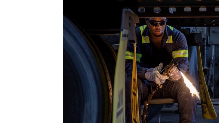 A mechanic in safety gear works under a truck trailer, holding a blowtorch while wearing gloves and protective glasses.