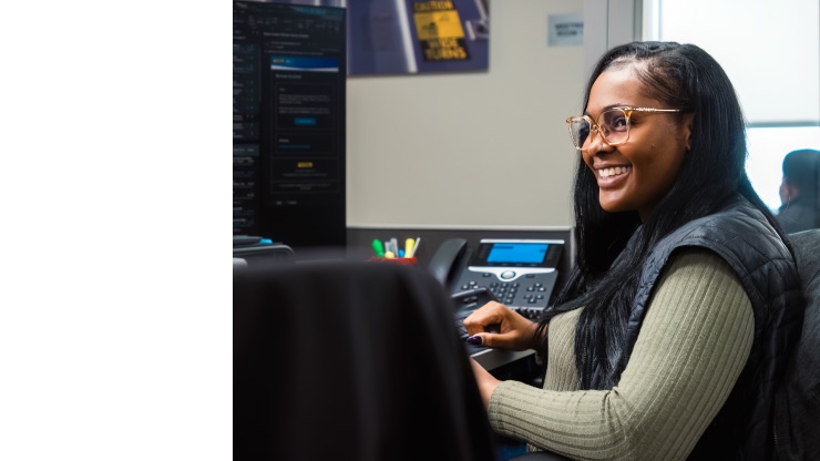 A woman wearing glasses and a green sweater, seated at a desk with a computer monitor, phone, and colorful office supplies, smiling in a professional office environment.