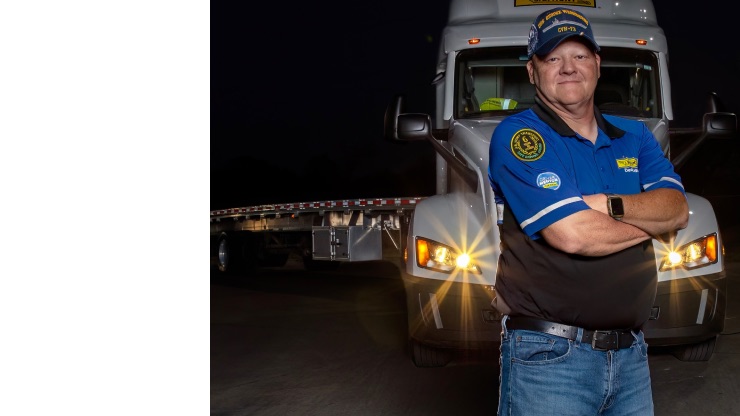 A professional truck driver wearing a branded uniform and cap stands confidently in front of a semi-truck at night, with headlights illuminated.