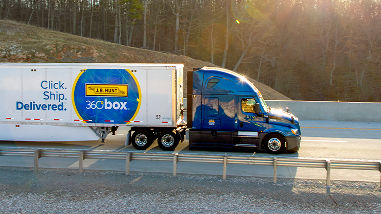 A blue semi truck hauls a white J.B. Hunt 360box trailer up a hill on a four lane highway